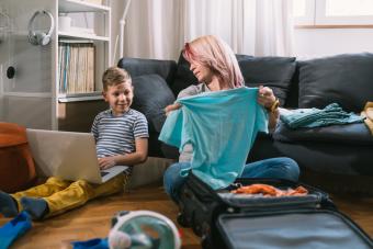 [image of a child and an adult sitting on the floor in front of a couch. The child is using a laptop computer and the adult is placing a shirt into a suitcase]